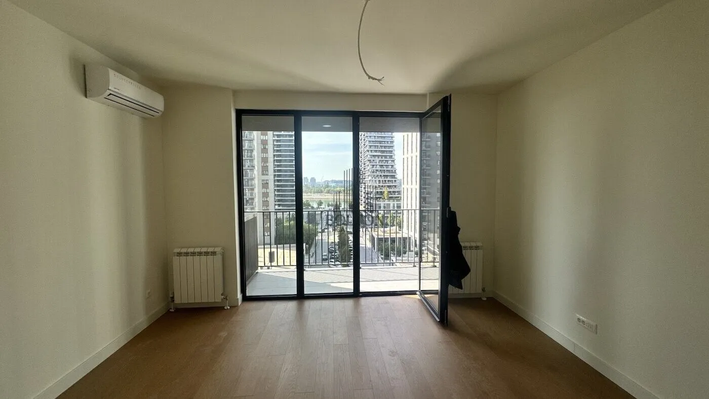 Prazna soba sa drvenim podom, klima uređajem i izlazom na balkon.