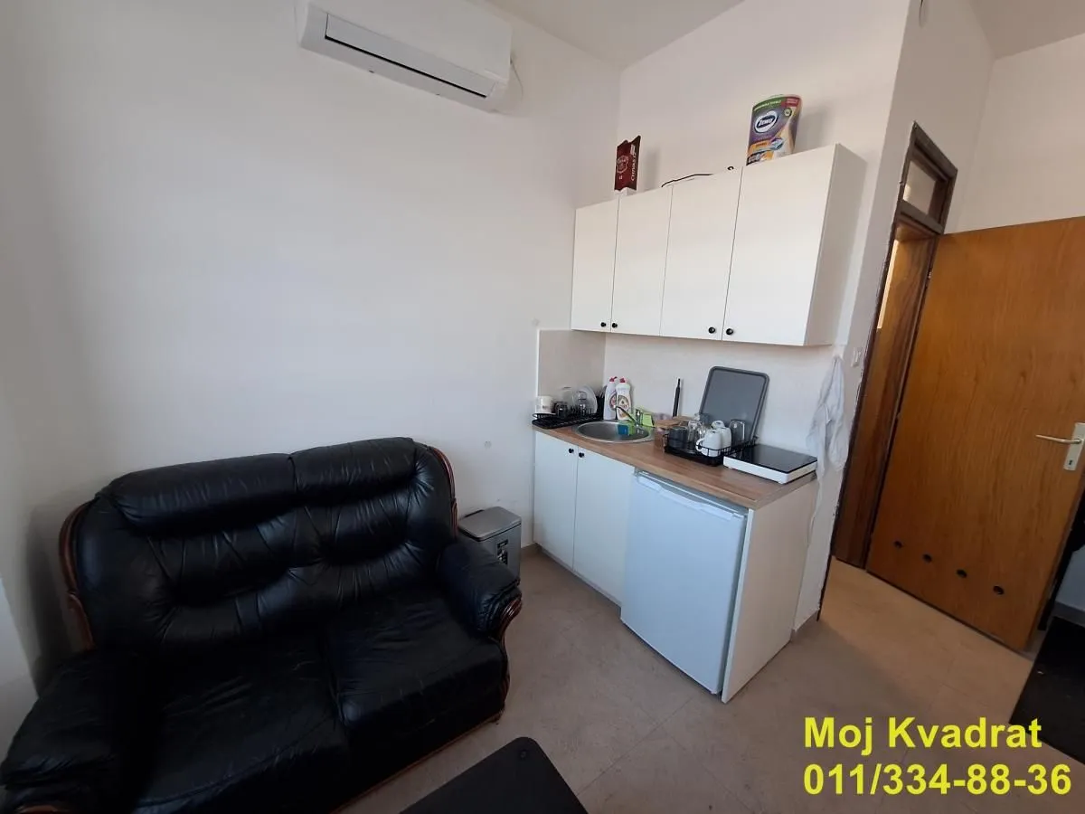 Small kitchen with white units and a black leather sofa in the same room.