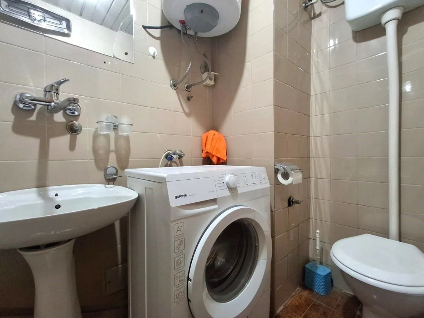 Manje kupatilo obloženo keramičkim pločicama sa umivaonikom, veš mašinom i toaletom.