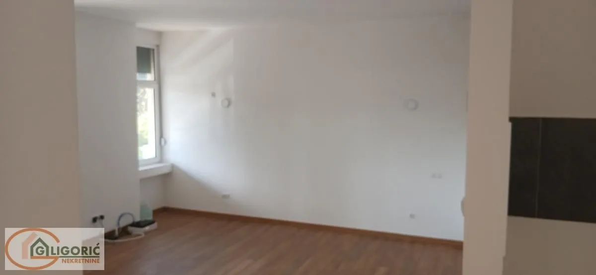 Prazna soba sa belim zidovima, drvenim podom i jednim prozorom.