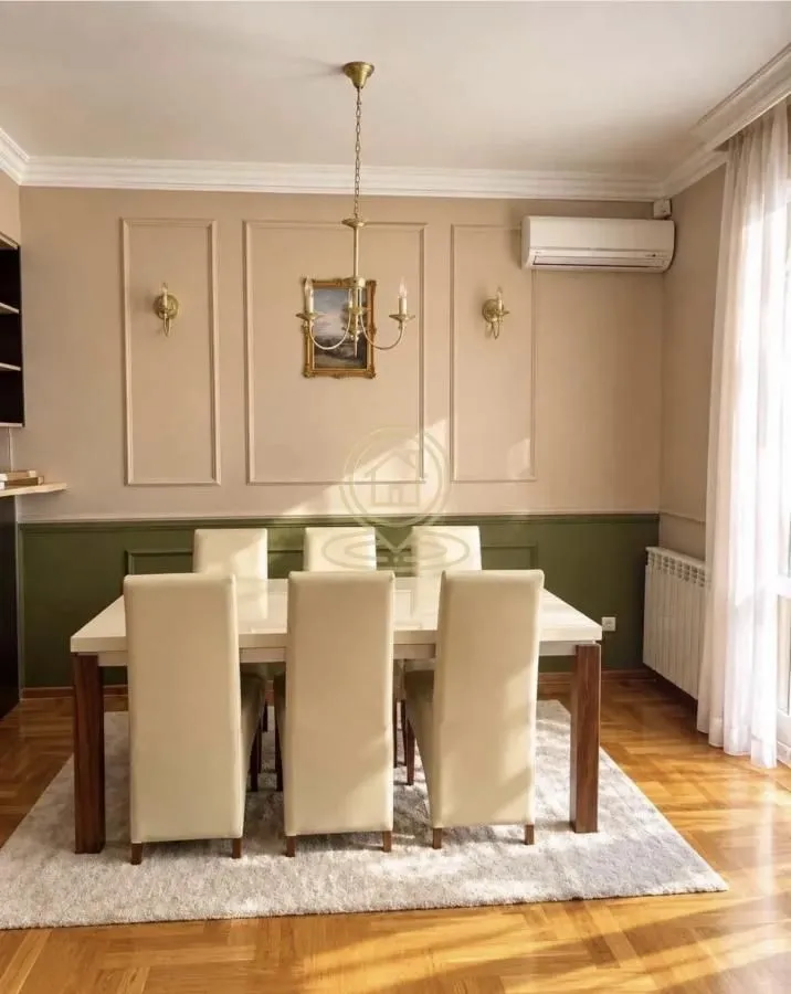 Trpezarija sa velikim stolom i stolicama, ukrašena zidnim lajsnama i lusterom.