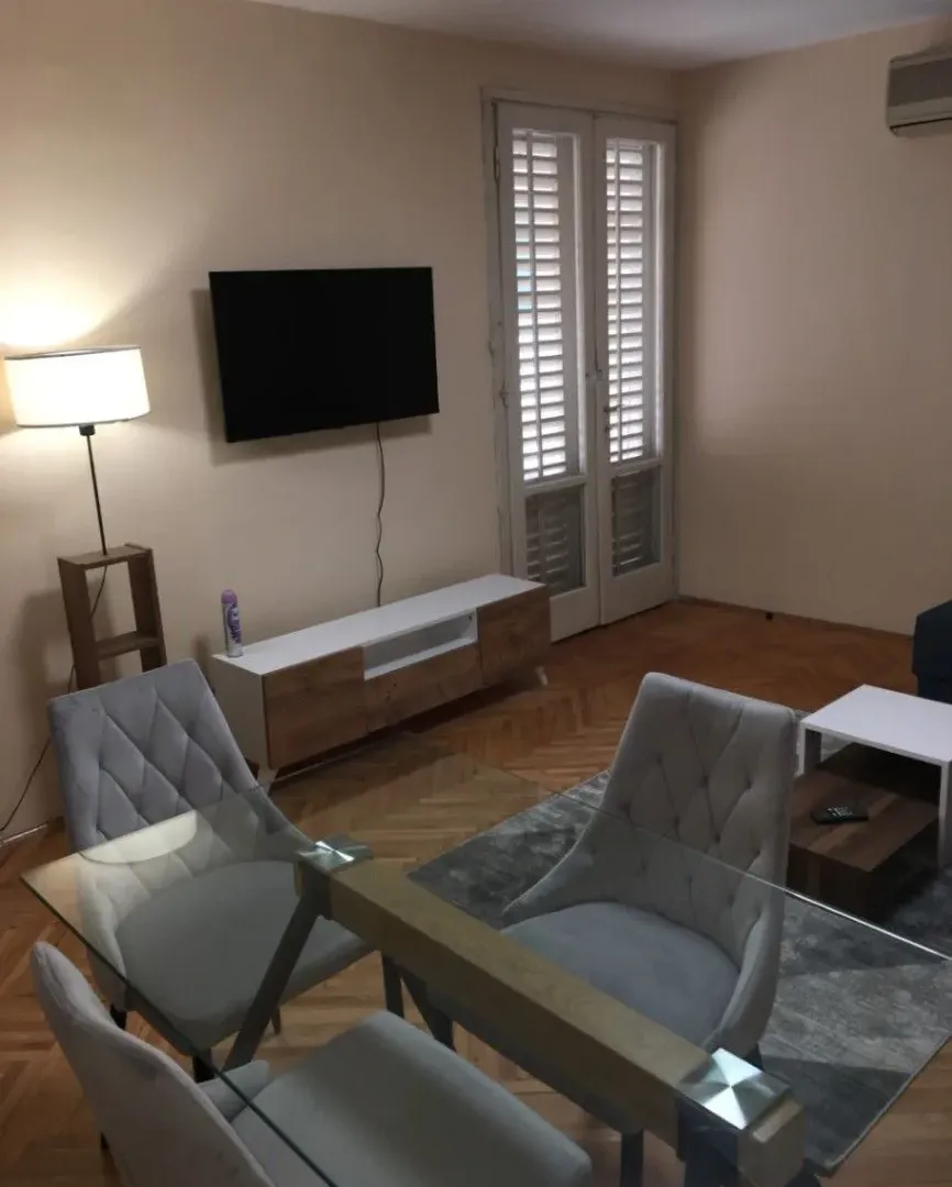 Dnevna soba sa trpezarijskim stolom i stolicama, TV-om i balkonskim vratima.