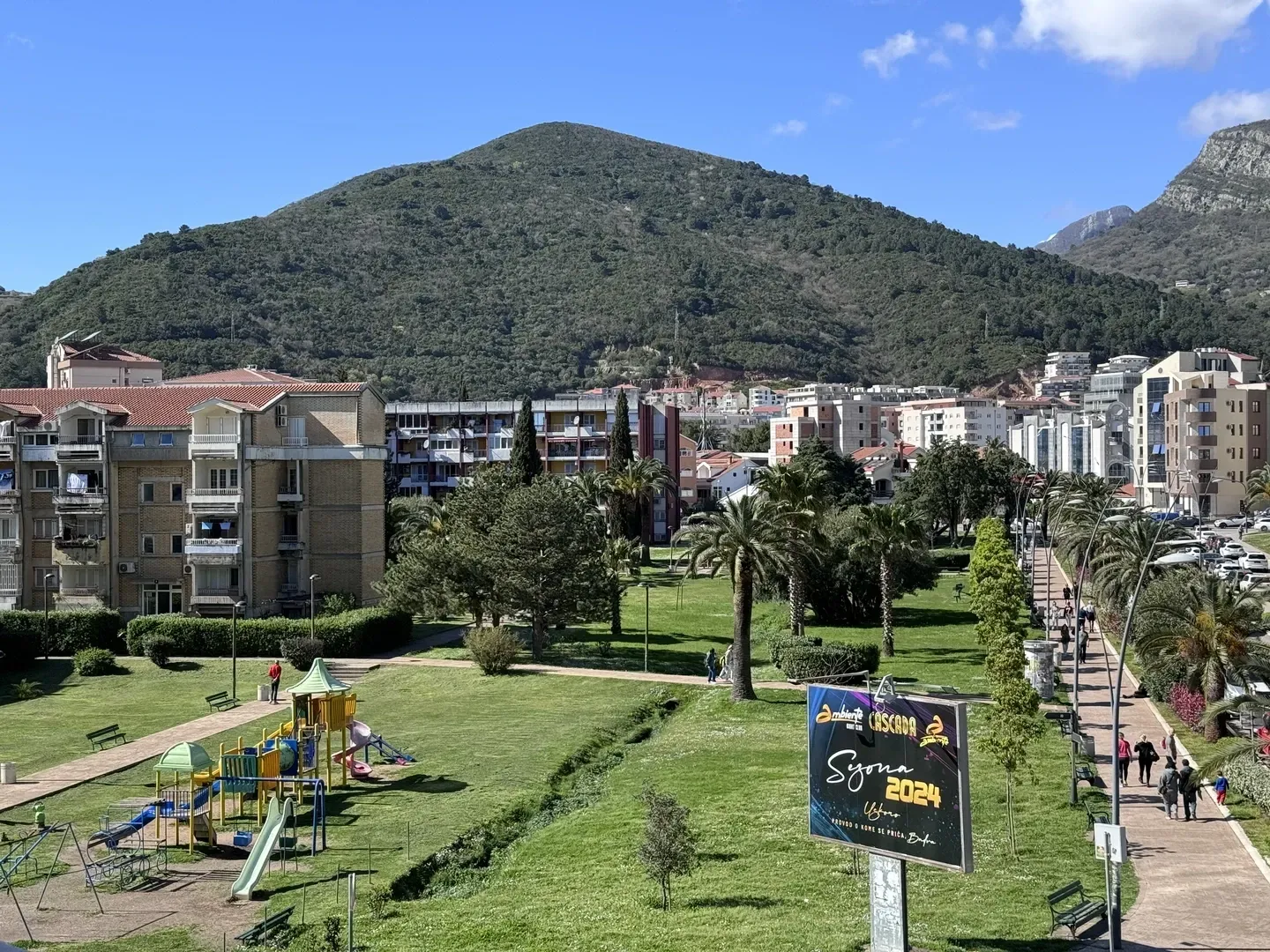 Pogled sa prozora na park, šetalište i brdo u pozadini.