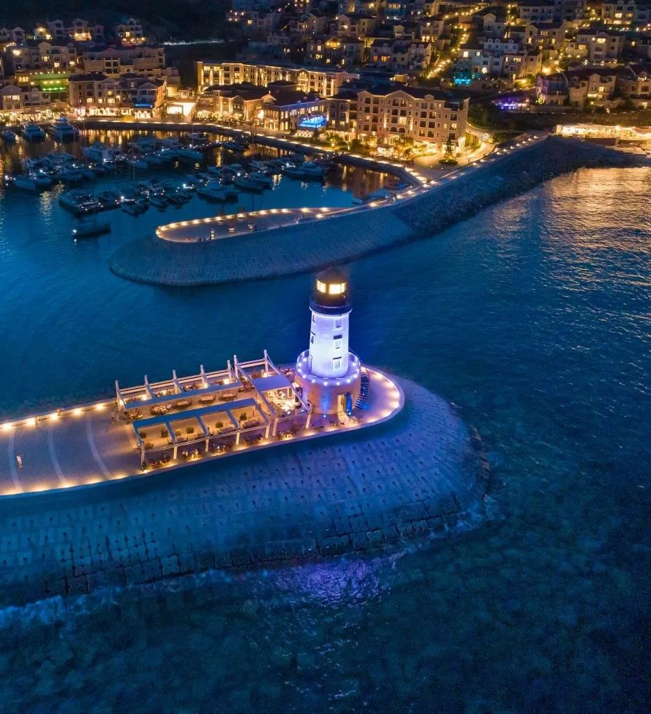 Noćni pogled iz vazduha na osvijetljeni svjetionik, marinu i obalu sa zgradama.