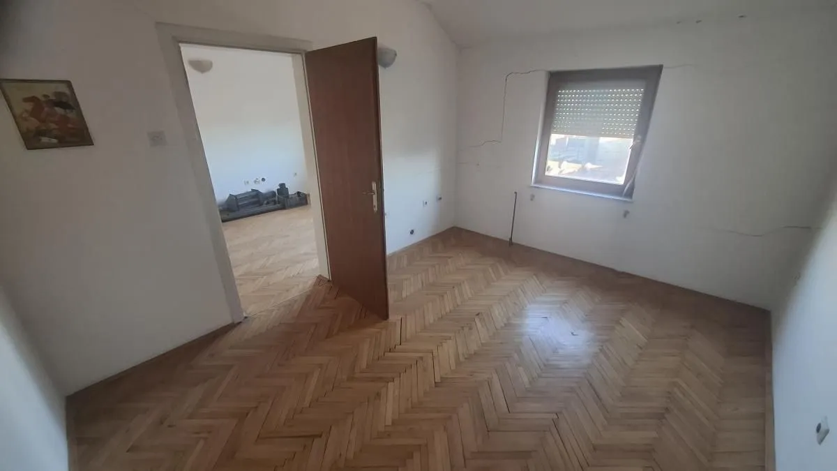 Prazna soba sa drvenim parketom i otvorenim vratima prema drugoj prostoriji.