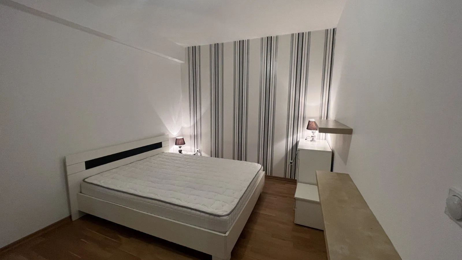 A simple bedroom with a double bed, striped wallpaper, and white furniture.