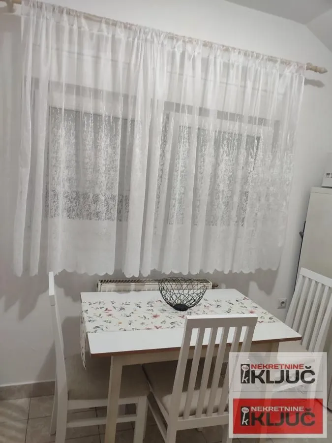 Trpezarijski sto sa belim stolicama ispred prozora sa zavesama.