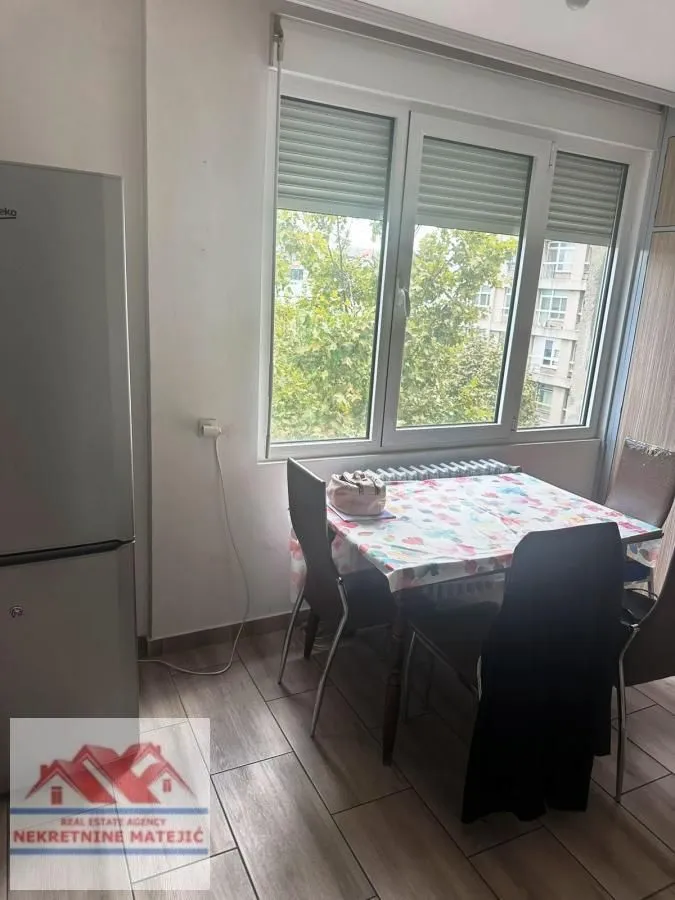 Trpezarijski deo sa stolom, stolicama i frižiderom pored prozora.