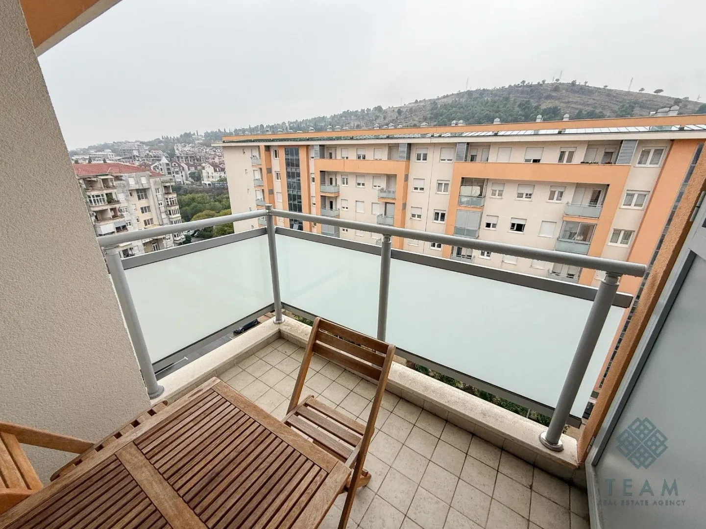 Manji popločani balkon sa drvenim stolom i stolicom i pogledom na naselje.
