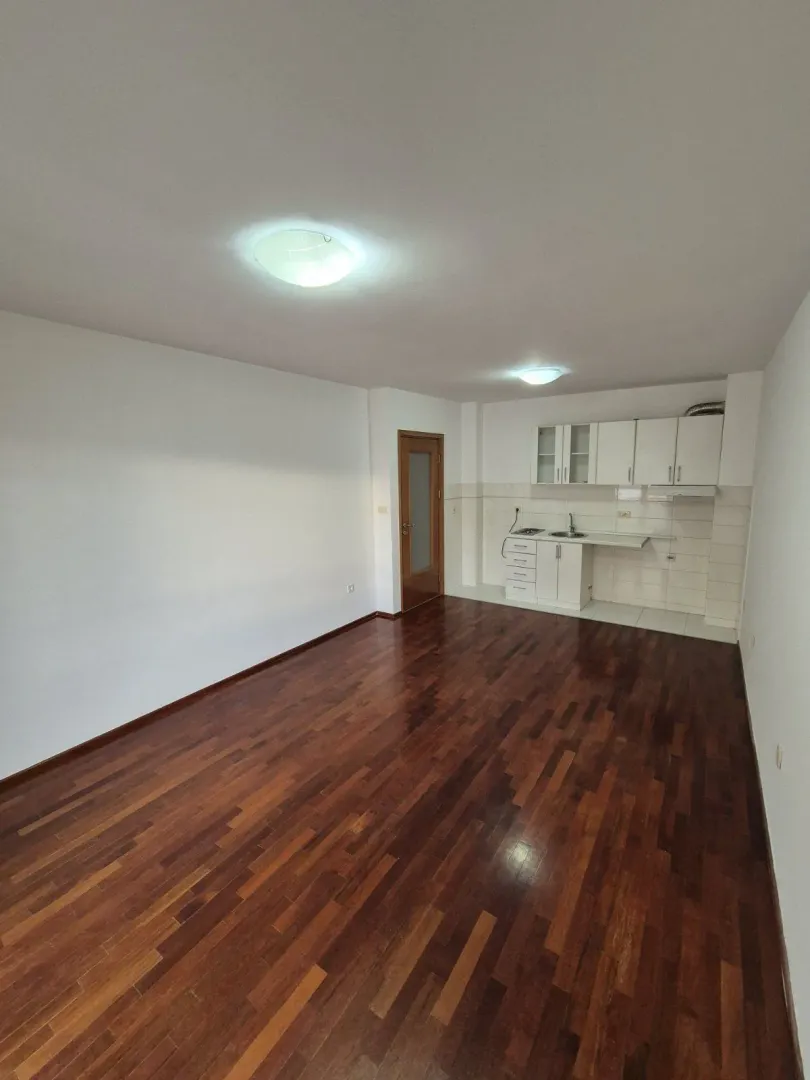 An empty room with dark parquet flooring and a small kitchen unit in the corner.