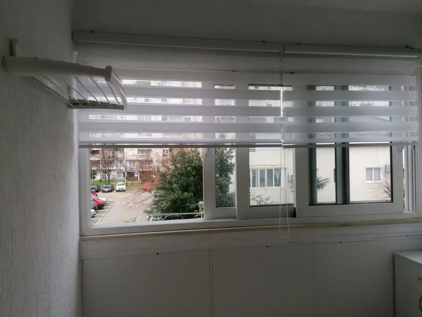 Pogled kroz prozor sa roletnama sa terase na okolne zgrade i parking.