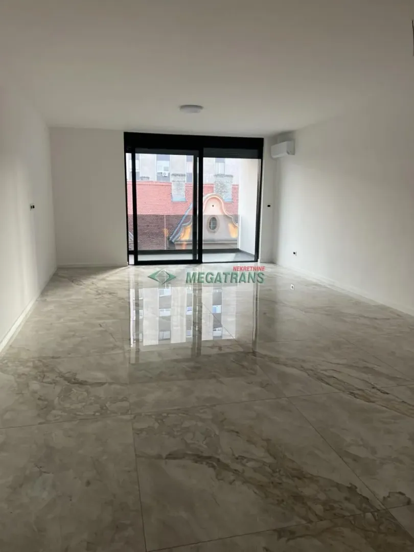 Prazna dnevna soba sa svetlim pločicama i velikim staklenim vratima koja izlaze na balkon.