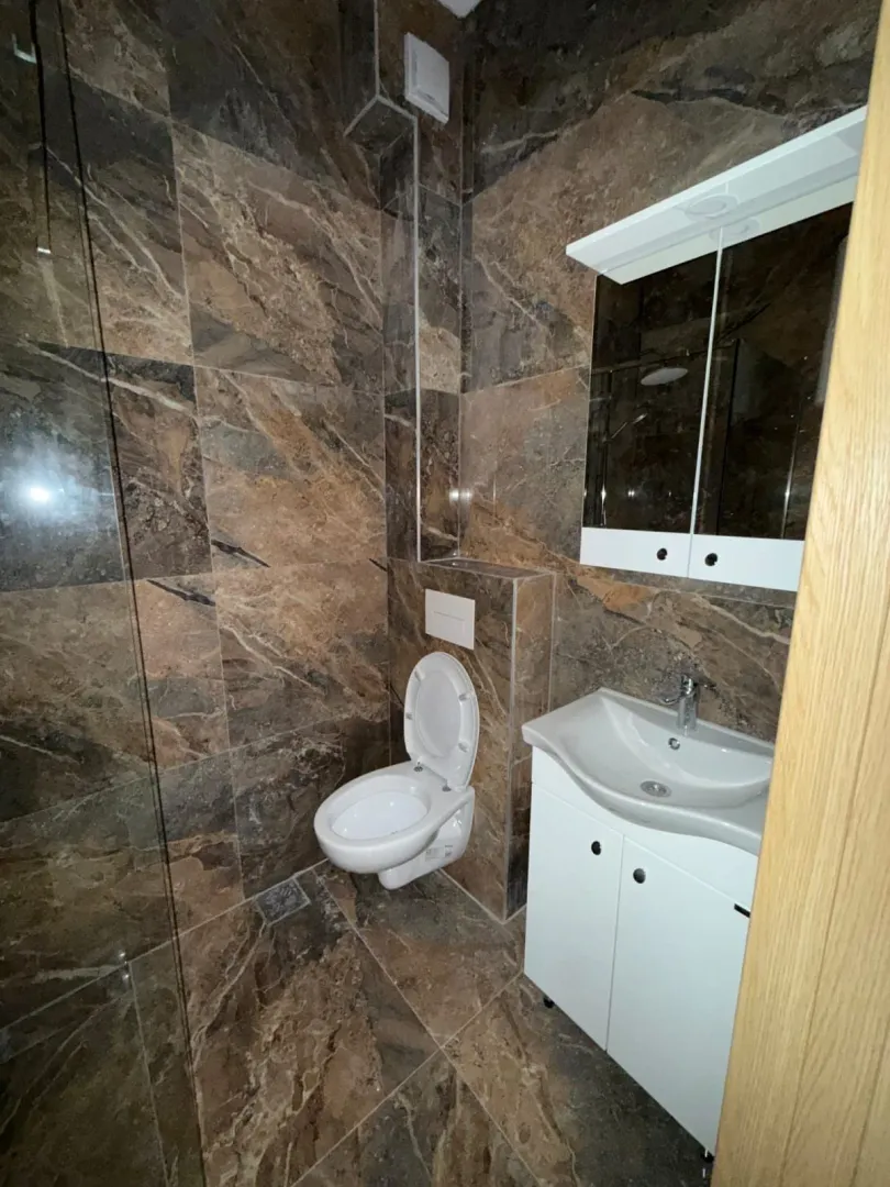 Manje kupatilo obloženo tamnim keramičkim pločicama sa sanitarijama i ogledalom.