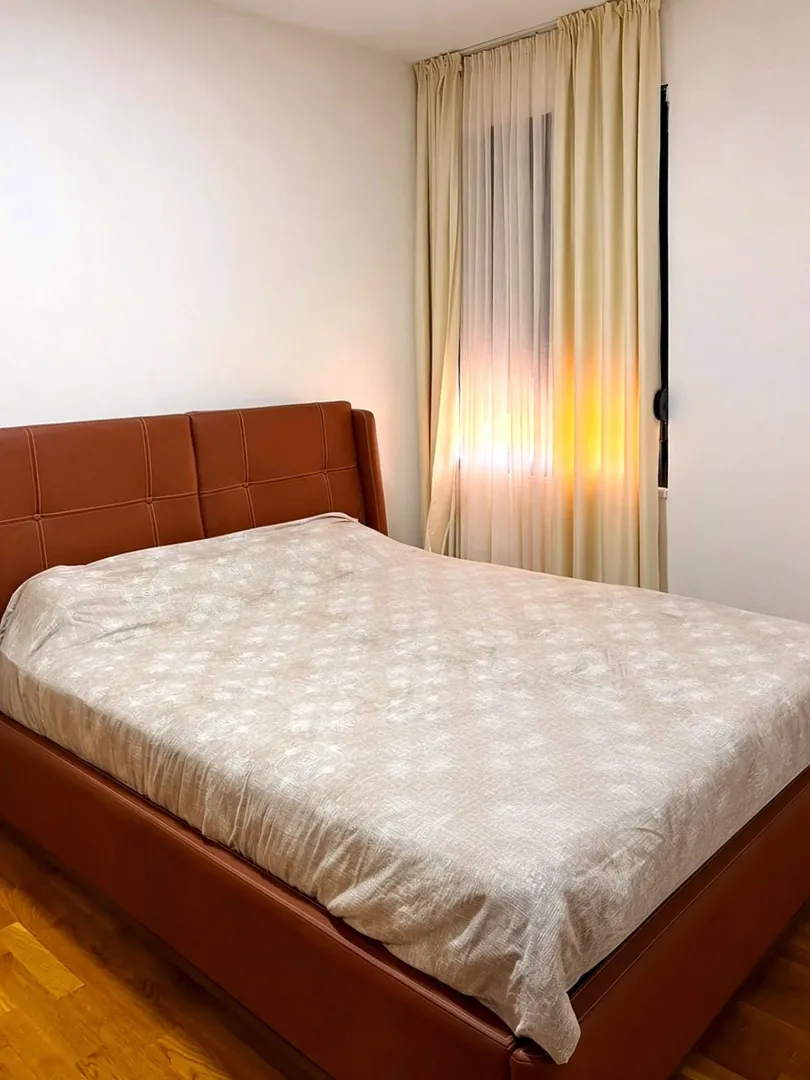 Spavaća soba sa bračnim krevetom sa smeđim tapaciranim uzglavljem i prozorom sa zavjesama.