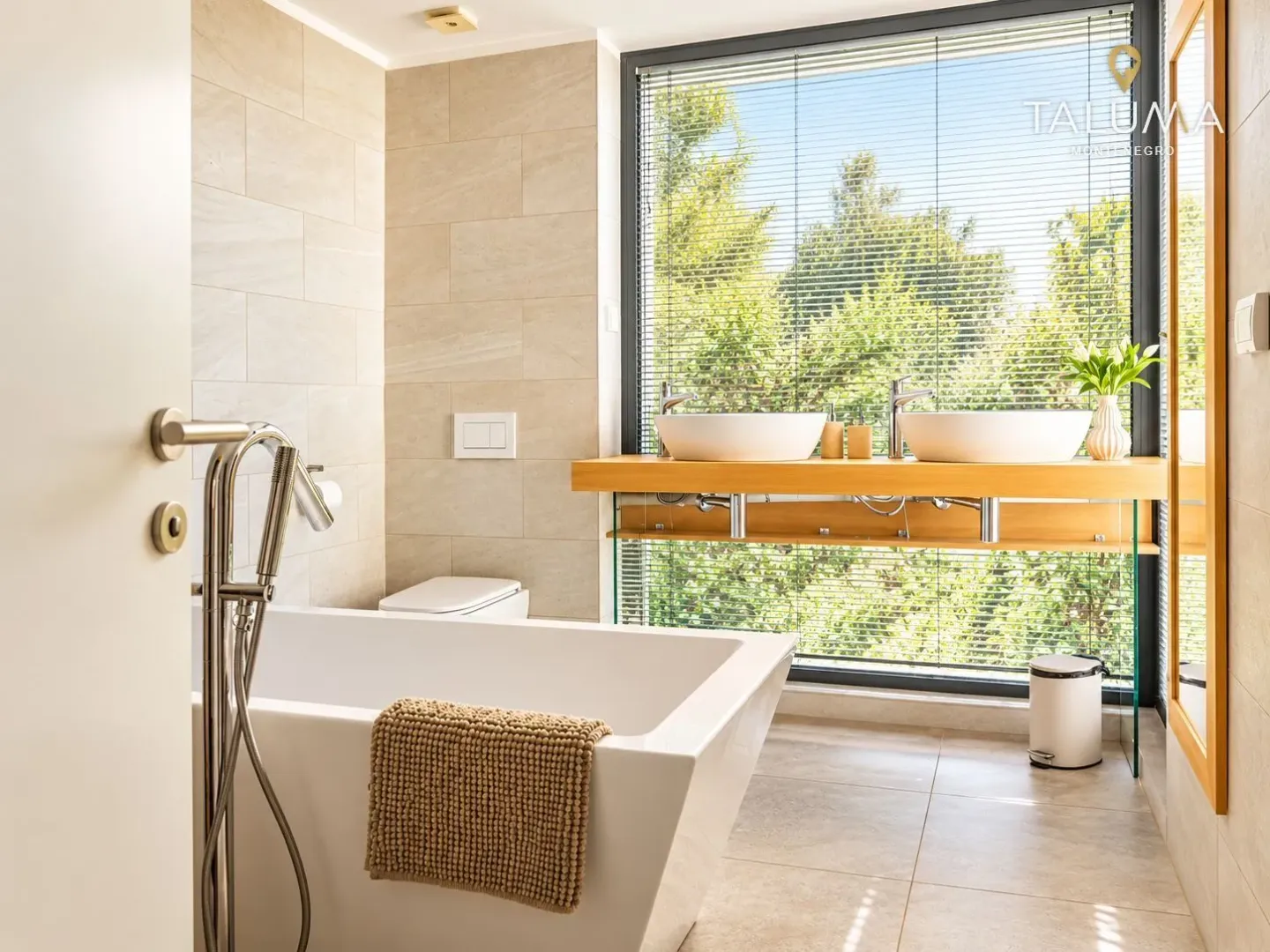 Modern bathroom with a bathtub, double sink, and a large window with a view of greenery.