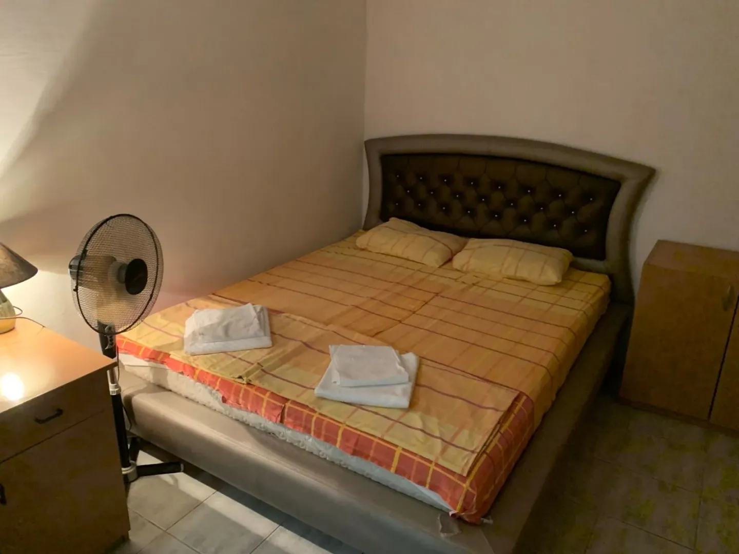Spavaća soba sa bračnim krevetom, noćnim stočićem i ventilatorom.