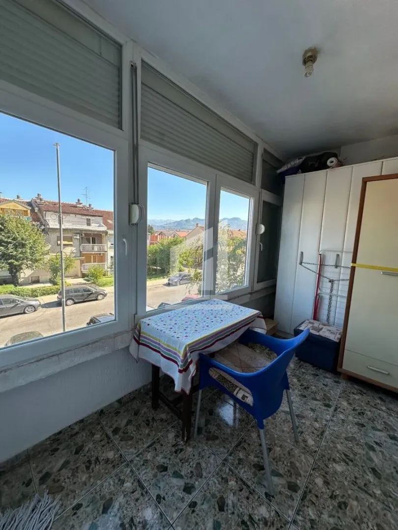 Zastakljeni balkon sa stolom, stolicom, ormarom i pogledom na okolinu.