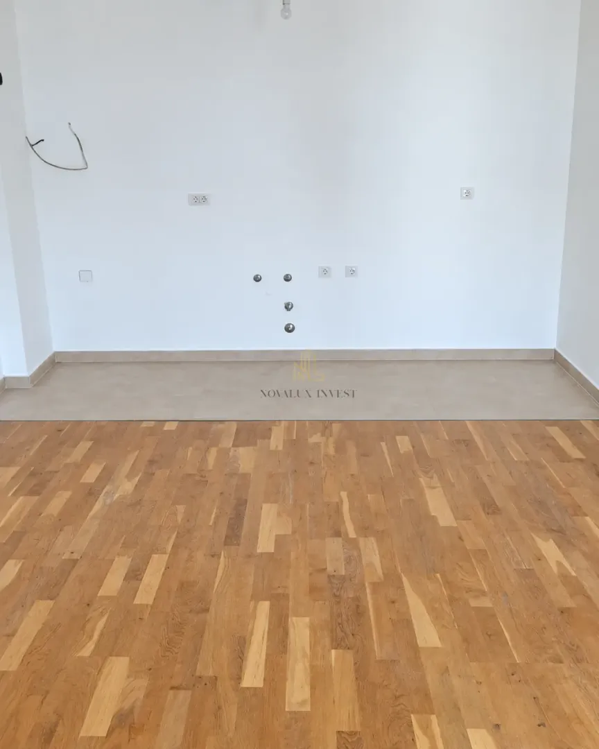 Empty kitchen space with utility connections on a white wall and wooden parquet flooring.