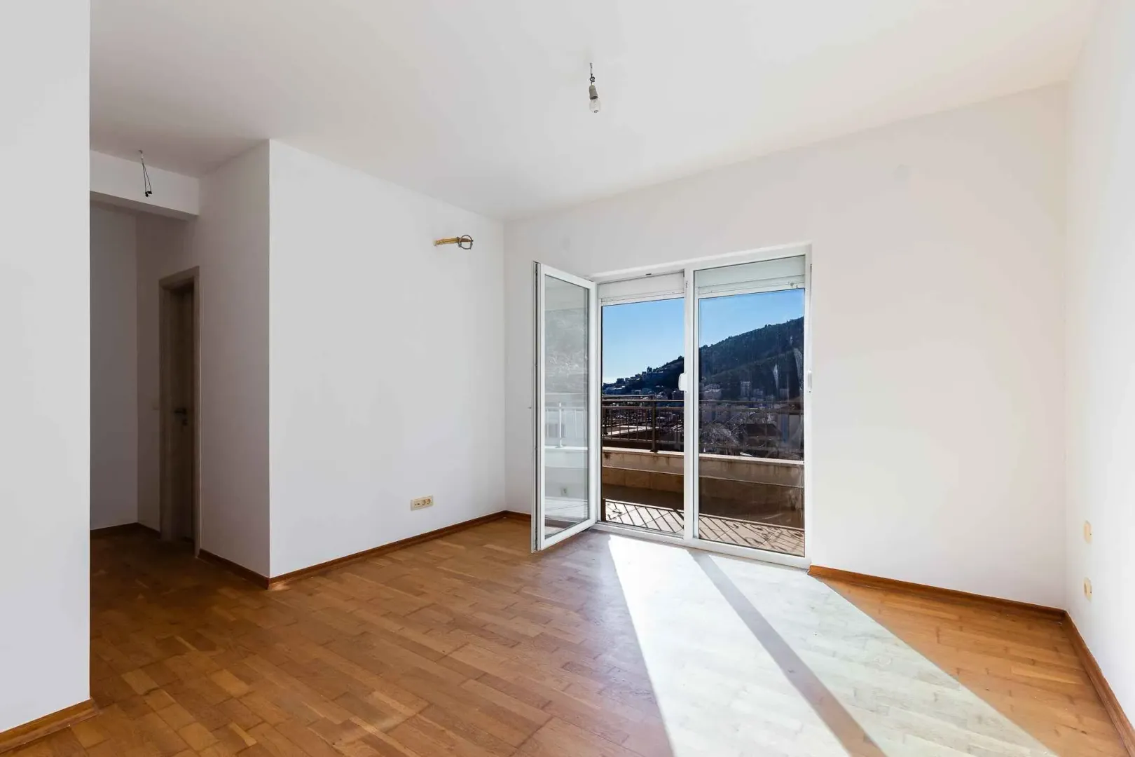 Prazna i svijetla prostorija sa parketom i izlazom na balkon.