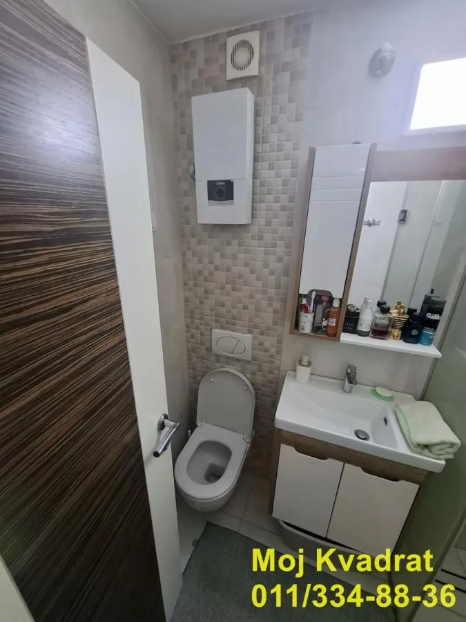 Manje kupatilo sa WC šoljom, umivaonikom, ogledalom i plinskim kotlom na zidu.