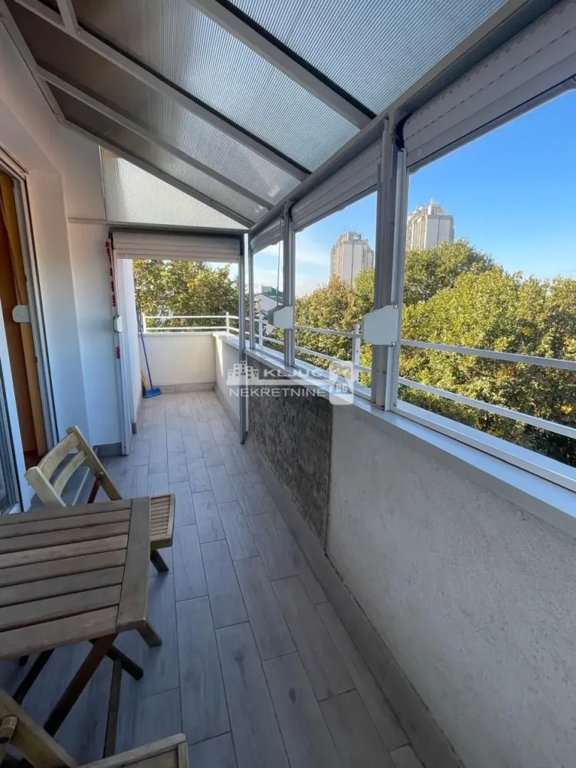 Zastakljeni balkon sa drvenim stolom i pogledom na zelenilo i zgrade.