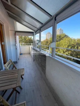 Zastakljeni balkon sa drvenim stolom i pogledom na zelenilo i zgrade. - image 10