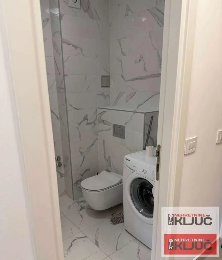 Pogled na kupatilo sa keramičkim pločicama, toaletom i veš mašinom.