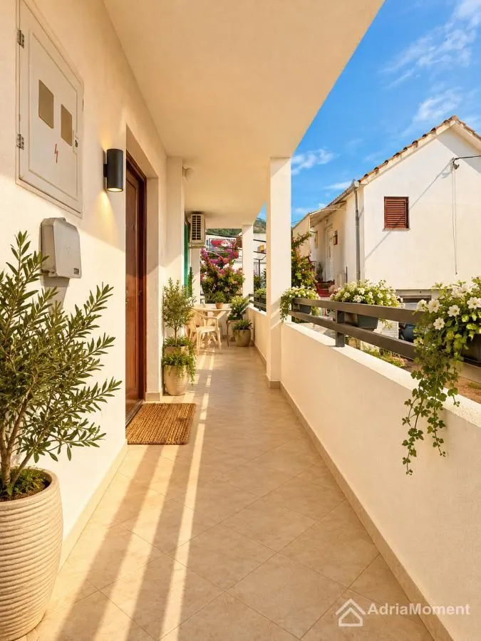 Dugački natkriveni balkon sa popločanim podom i saksijama sa cvijećem.
