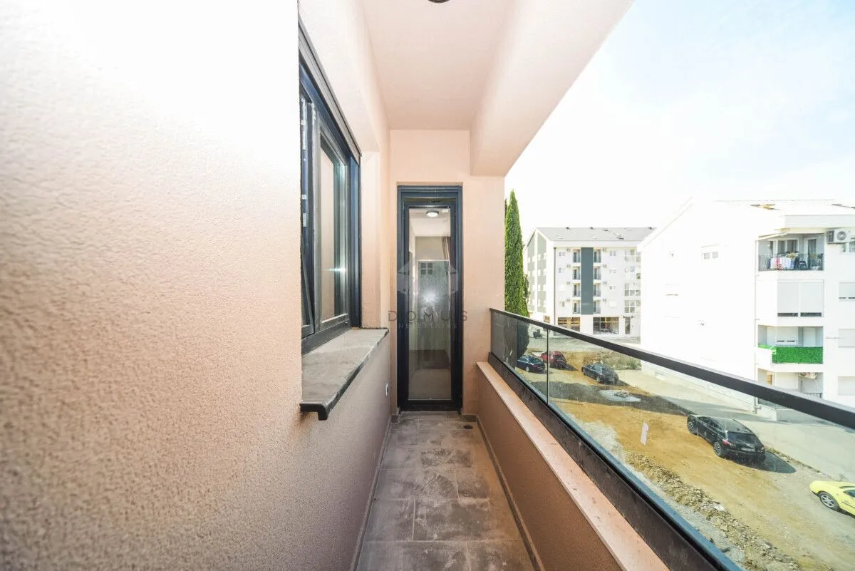 Dugačak balkon sa staklenom ogradom i pogledom na okolne zgrade i parking.