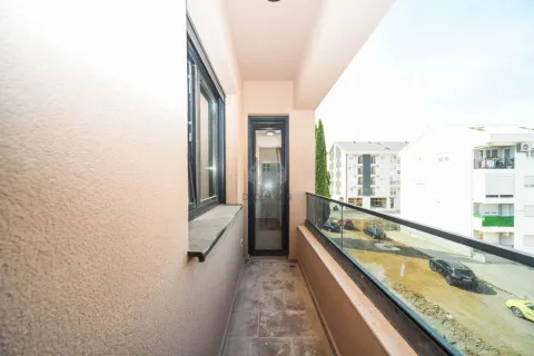 Dugačak balkon sa staklenom ogradom i pogledom na okolne zgrade i parking. - image 36