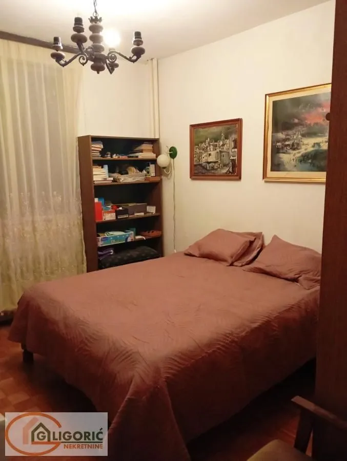 Nameštena spavaća soba sa bračnim krevetom, police za knjige i slikama na zidu.