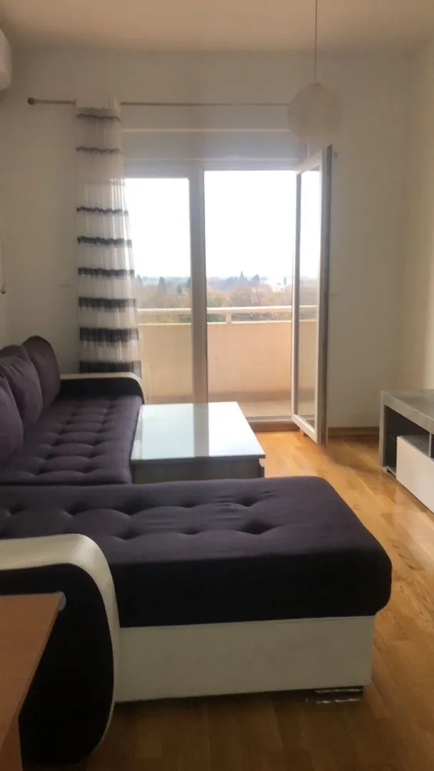 Svijetla dnevna soba sa tamnom ugaonom garniturom i izlazom na balkon.