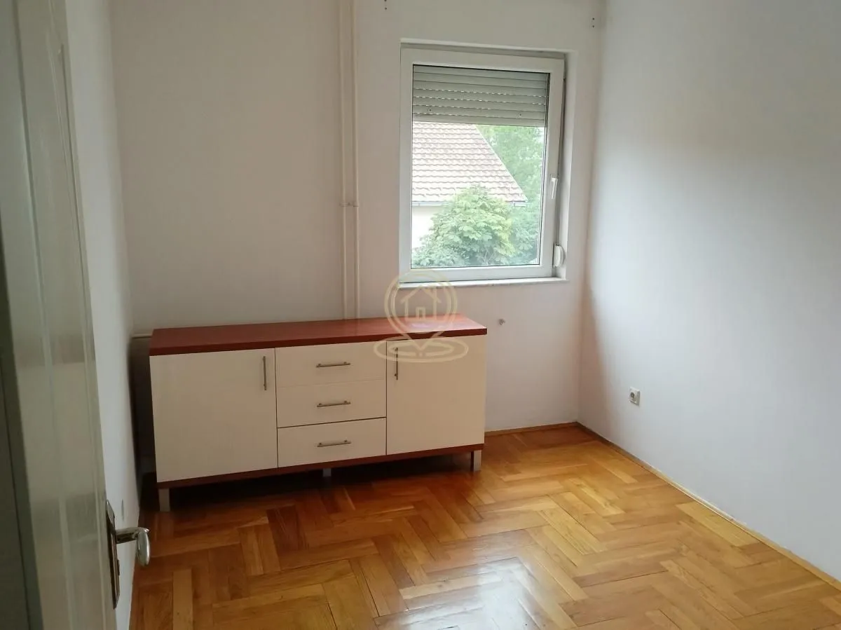 Manja soba sa drvenim parketom, komodom i prozorom.