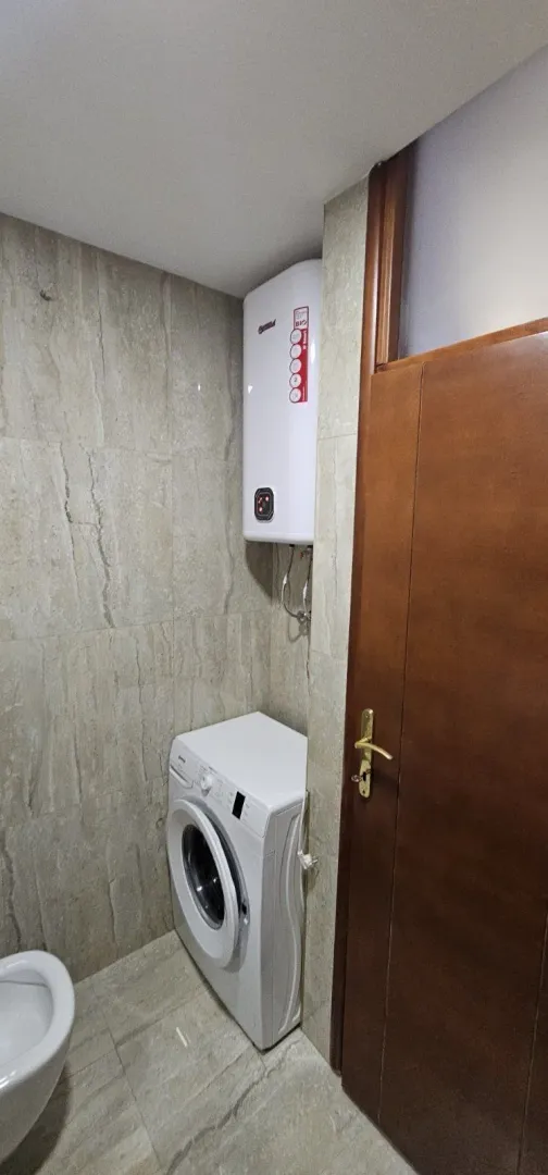 Dio kupatila sa veš mašinom i bojlerom pored drvenih vrata.