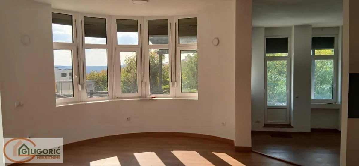 Prazna dnevna soba sa velikim polukružnim prozorima i drvenim podom.