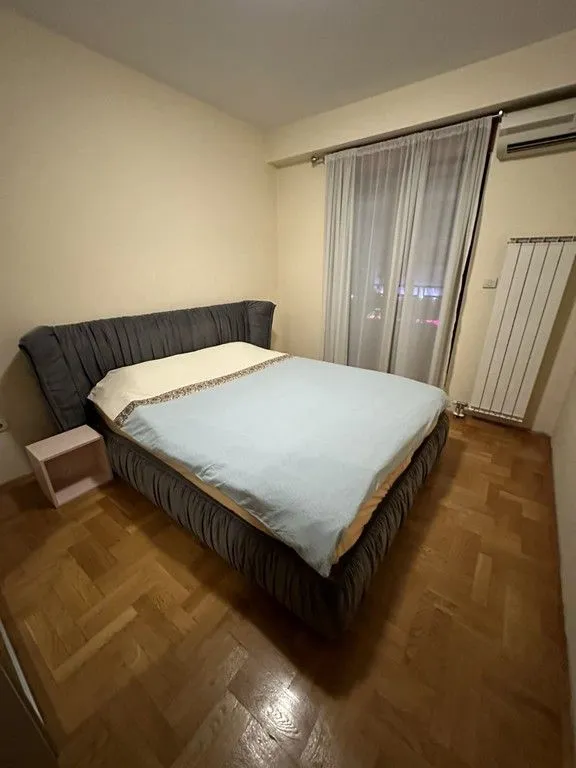 Spavaća soba sa bračnim krevetom, drvenim parketom i prozorom sa zavjesama.