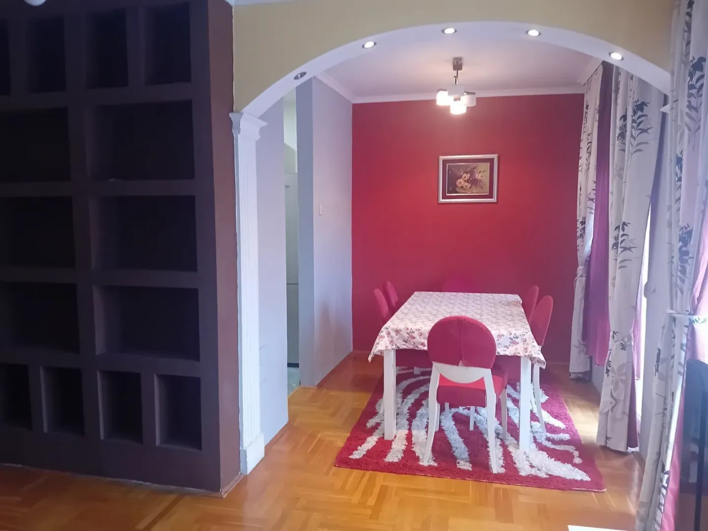 Trpezarijski prostor sa crvenim akcentnim zidom, stolom i stolicama ispod ukrasnog luka.