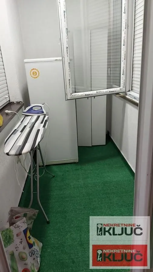 Zastakljeni balkon sa veštačkom travom, zamrzivačem i daskom za peglanje.