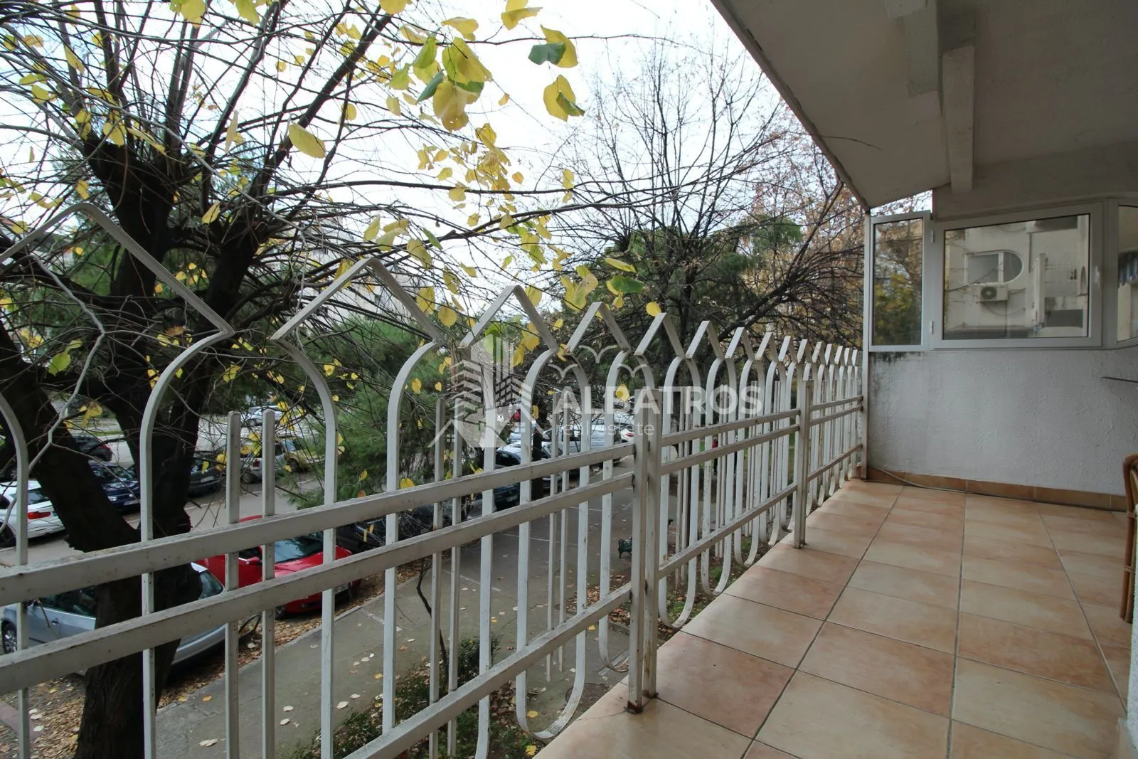 Natkriveni balkon sa popločanim podom i metalnom ogradom, sa pogledom na ulicu i drveće.