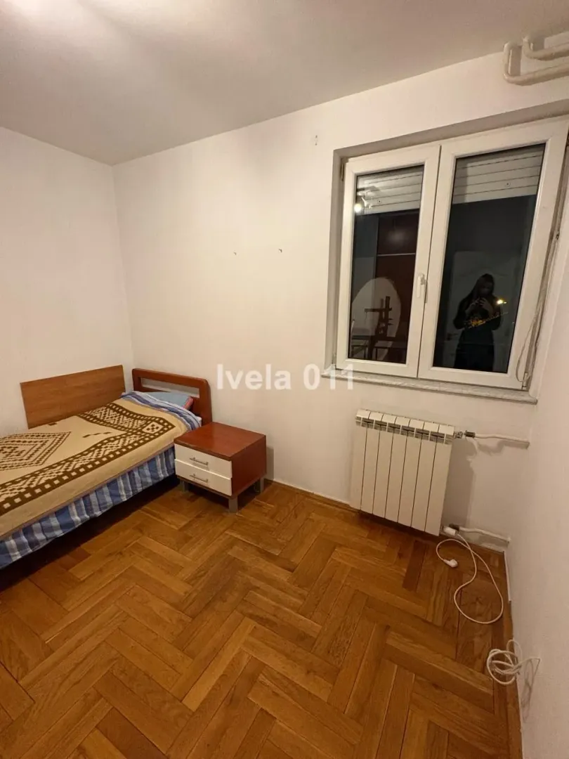 Manja spavaća soba sa krevetom, noćnim stočićem, prozorom i radijatorom.