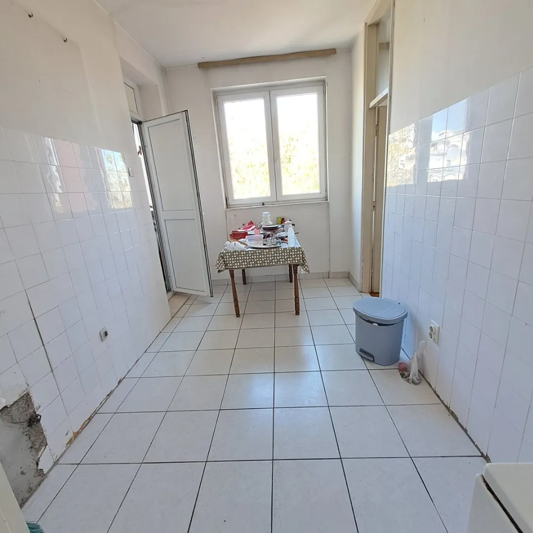 Prazna kuhinja sa bijelim pločicama na zidovima i podu, sa trpezarijskim stolom kod prozora.
