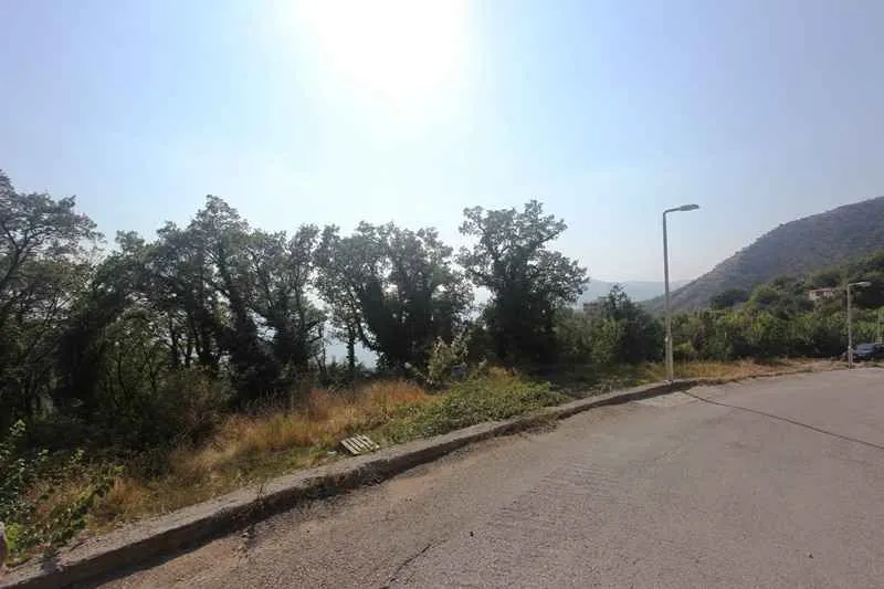Pogled na građevinski plac pored asfaltnog puta sa drvećem i brdima u pozadini.
