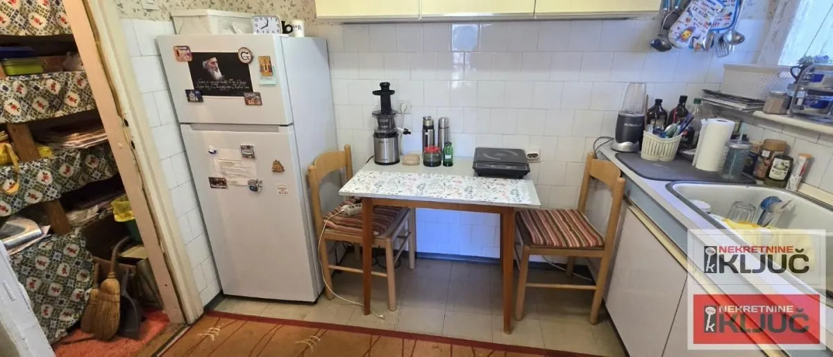 Manja kuhinja sa frižiderom, sudoperom i trpezarijskim stolom sa dve stolice.