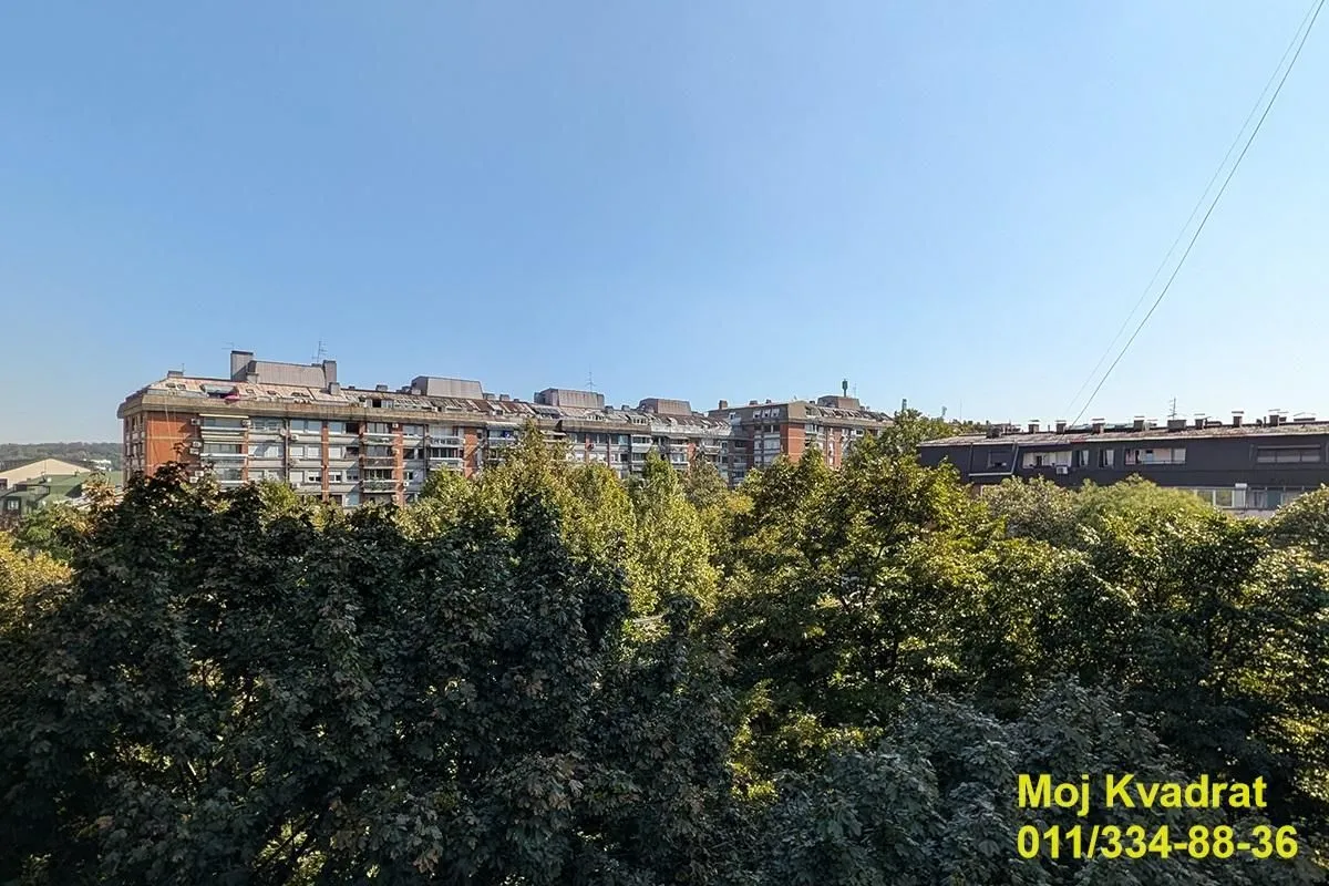 Pogled sa prozora na okolne zgrade i zelene krošnje drveća.