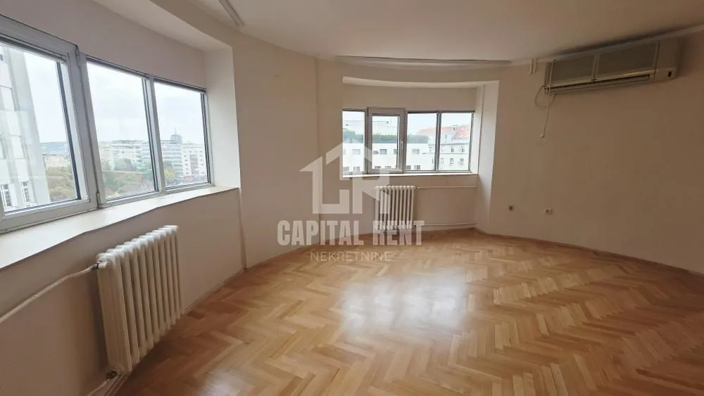 Empty living room with parquet flooring, radiators, and large windows.