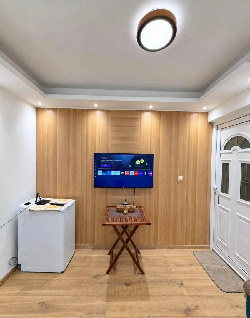 Dnevni boravak sa drvenim zidnim panelima, televizorom i ulaznim vratima.