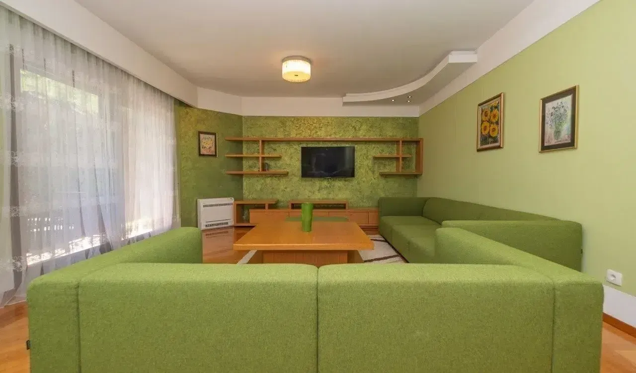 Dnevna soba sa zelenom garniturom, drvenim stolićem i televizorom na zidu.