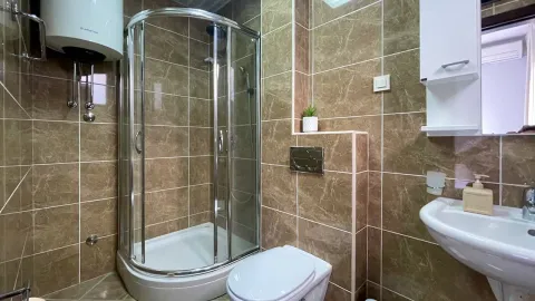 Kupatilo obloženo smeđim pločicama sa tuš kabinom, WC šoljom i umivaonikom. - image 24