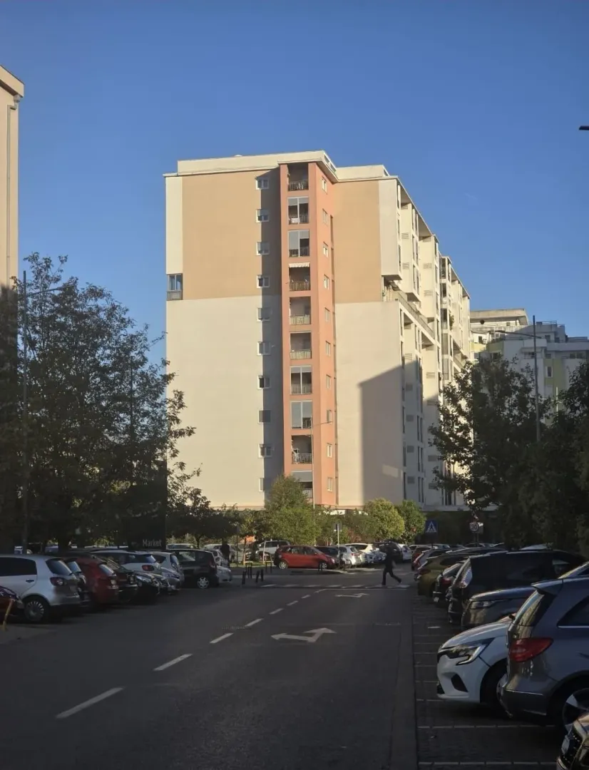 Pogled na stambenu zgradu sa parkingom i ulicom u prvom planu.