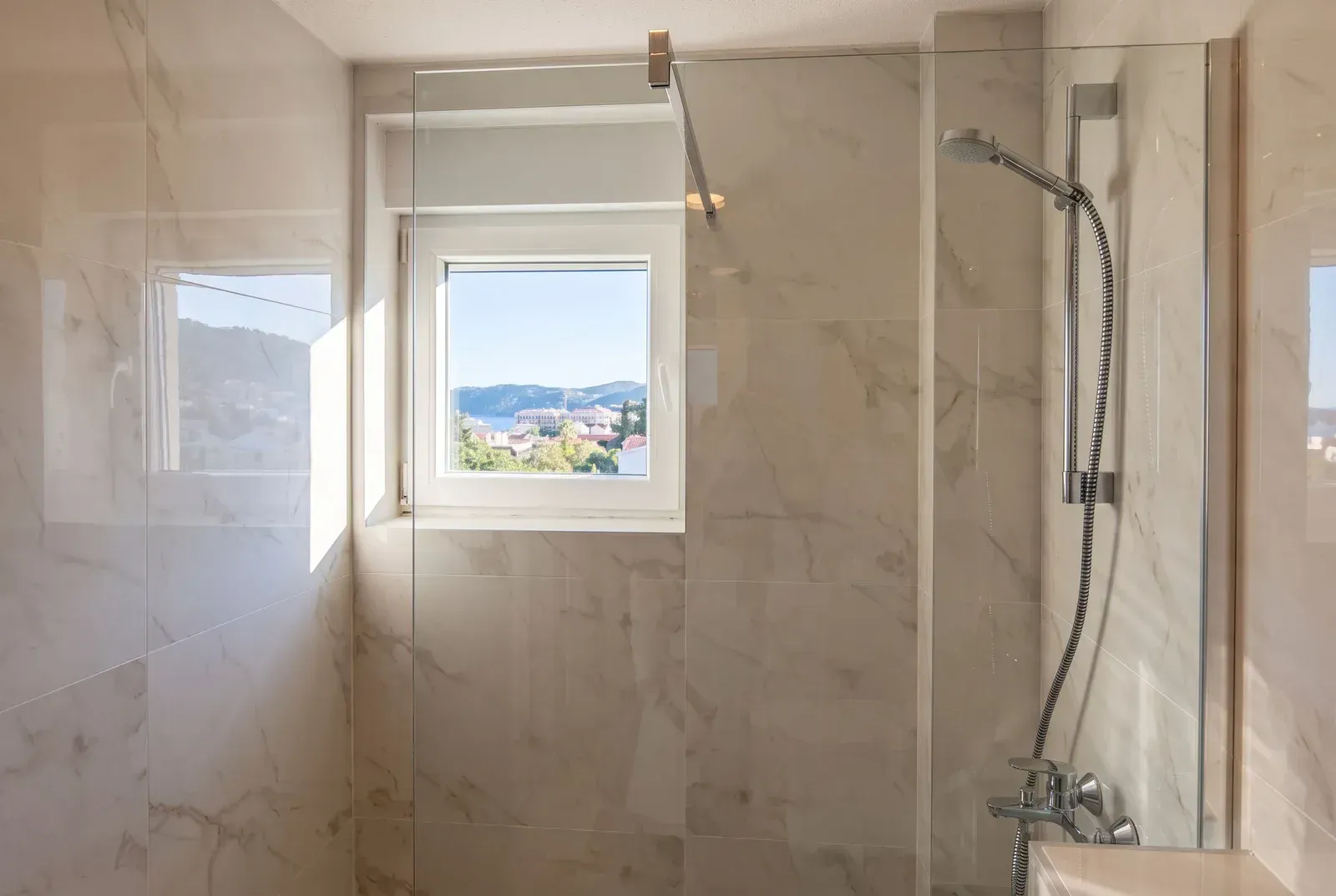 Modern walk-in shower with marble tiles and a view through the window.